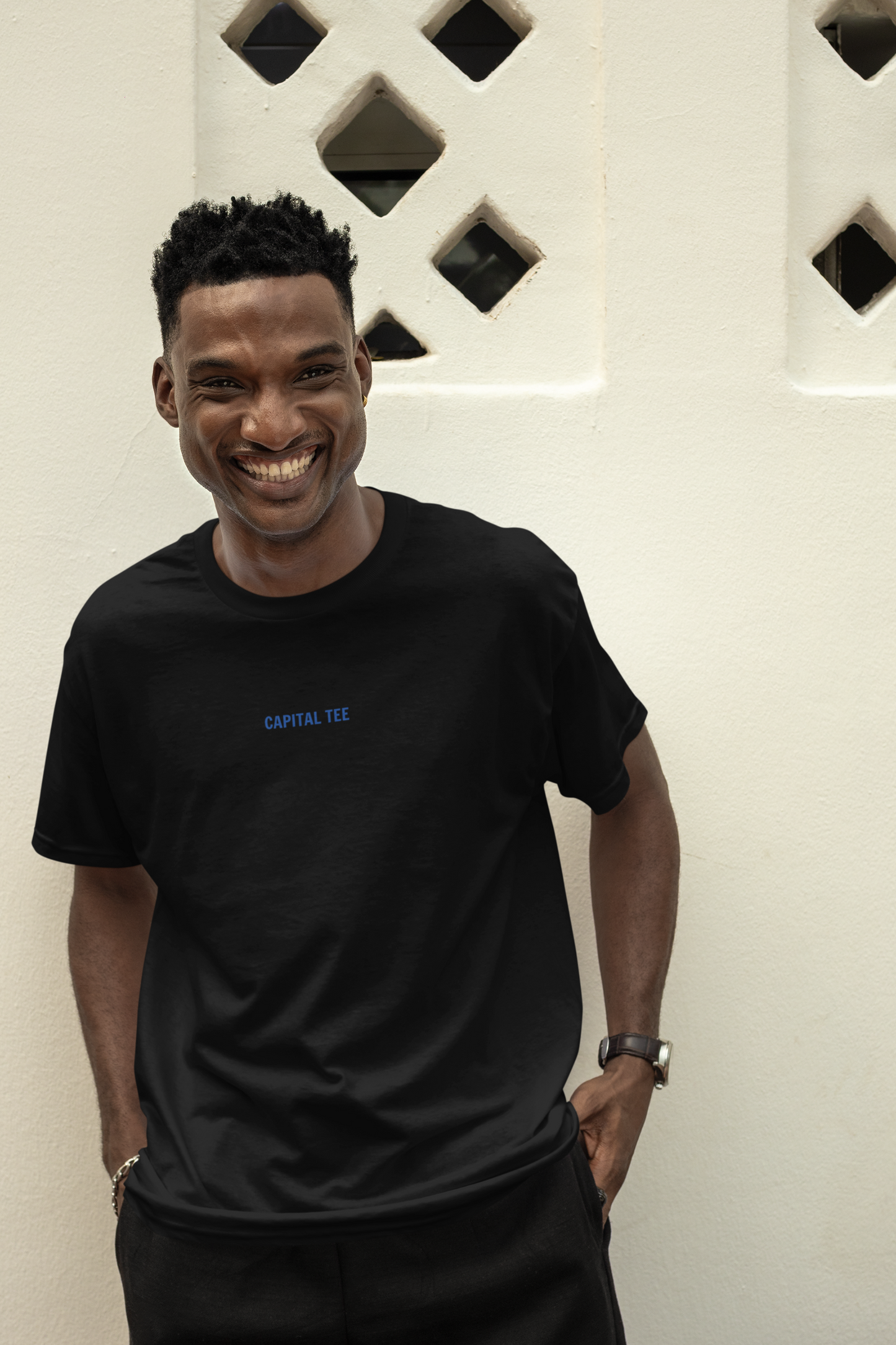 A man smiling while wearing a black T-shirt that says 'CAPITAL TEE' in blue text on the front.